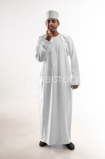 الاشارة إلى الفم بإيماءات الانزعاج والألم, مشاكل التهاب اللثة, طلب العلاج والمساعدة, الحاجة لزيارة طبيب الأسنان, بورتريه مقرب لشاب عربي خليجي عماني يرتدي الدشداشة و الكمه يضع يده على فمه, صورة الجسم بالطول الكامل, خلفية بيضاء