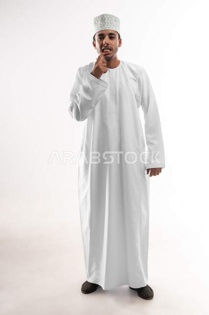 الاشارة إلى الفم بإيماءات الانزعاج والألم, مشاكل التهاب اللثة, طلب العلاج والمساعدة, الحاجة لزيارة طبيب الأسنان, بورتريه مقرب لشاب عربي خليجي عماني يرتدي الدشداشة و الكمه ينظر الى الكاميرا يضع يده على فمه, صورة الجسم بالطول الكامل, خلفية بيضاء
