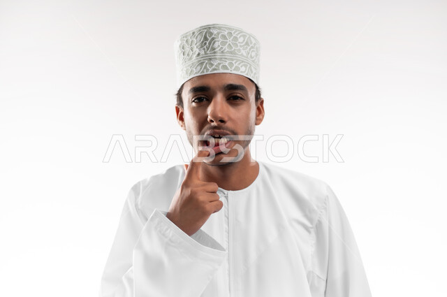 الاشارة إلى الفم بإيماءات الانزعاج والألم, مشاكل التهاب اللثة, طلب العلاج والمساعدة, الحاجة لزيارة طبيب الأسنان, بورتريه مقرب لشاب عربي خليجي عماني يرتدي الدشداشة و الكمه يضع يده على فمه, خلفية بيضاء