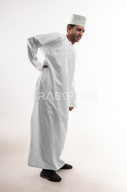 الأوجاع والآلام العصبية، طلب المساعده والحاجه للعلاج، التهاب العضلات والمفاصل، بورتريه لشاب عربي خليجي عماني يرتدي الدشداشة و الكمه يضع يده على ظهره بايماءات الألم والتعب والأرهاق، حالة اسعافية طارئة، صورة الجسم الطول الكامل, خلفية بيضاء