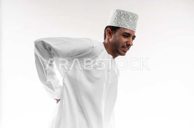 الأوجاع والآلام العصبية، طلب المساعده والحاجه للعلاج، التهاب العضلات والمفاصل، بورتريه مقرب لشاب عربي خليجي عماني يرتدي الدشداشة و الكمه يضع يده على ظهره بايماءات الألم والتعب والأرهاق، حالة اسعافية طارئة، خلفية بيضاء