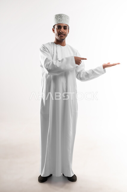 الامساك بعنصر ما بإيماءات الاعجاب، بورتريه لشاب عربي خليجي عماني يرتدي الدشداشة و الكمه ينظر الى الكاميرا يشير بإصبع السبابة ليده الأخرى، مفهوم الانتباه والتركيز، صورة الجسم بالطول الكامل, خلفية بيضاء