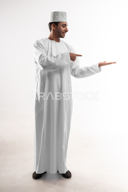 الامساك بعنصر ما بإيماءات الاعجاب، بورتريه لشاب عربي خليجي عماني يرتدي الدشداشة و الكمه يشير بإصبع السبابة ليده الأخرى، مفهوم الانتباه والتركيز، صورة الجسم بالطول الكامل, خلفية بيضاء