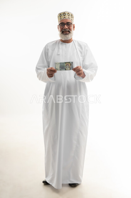 The official currency in circulation in the markets of the Sultanate of Oman, display of the new issue of Omani paper money, portrait of an old Arab Gulf Omani man wearing a dishdasha, a sleeve and eyeglasses, standing and holding a twenty riyal banknote with gestures of pleasure, full body, white background