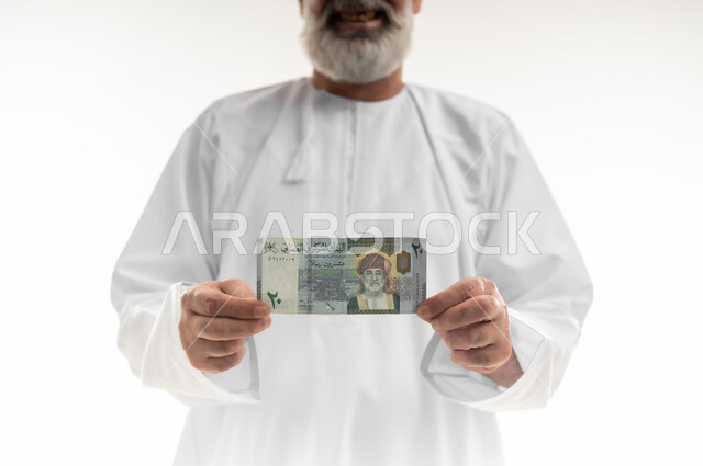 عرض الإصدار الجديد للنقود الورقية العمانية، العملة الرسمية المتداولة في أسواق سلطنة عمان،  بورتريه مقرب لرجل شايب عربي خليجي عماني مبتسم يرتدي الدشداشة التقليدية يقف ويمسك ورقة نقدية من فئة العشرين ريال بإيماءات السرور، خلفية بيضاء