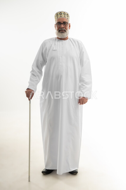 استخدام وسائل المساعدة في المشي والتحرك، الاعتماد على العصا الطبية لتحسين جودة حياة كبار السن، بورتريه لرجل شايب عربي خليجي عماني يرتدي الدشداشة والكمه العمانية يستند على عكازه من أجل التنقل من مكان إلى آخر، الجسم بالطول الكامل، خلفية بيضاء