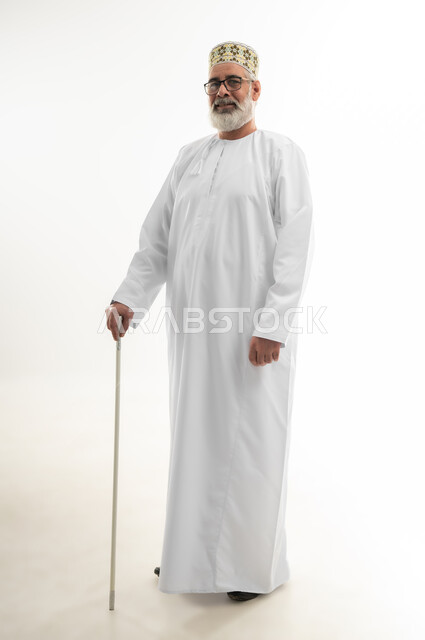 الاعتماد على العصا الطبية لتحسين جودة حياة كبار السن، استخدام وسائل المساعدة في المشي والتحرك، بورتريه لرجل شايب عربي خليجي عماني يرتدي الدشداشة والكمه العمانية يستند على عكازه من أجل التنقل من مكان إلى آخر، الجسم بالطول الكامل، خلفية بيضاء