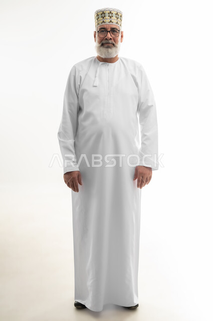 الثقة بالنفس والوقوف بشكل مستقيم، الاهتمام بالمظهر الخارجي، مفهوم الرجولة والوقار، بورتريه لرجل كبير بالسن عربي خليجي عماني يرتدي الدشداشة التقليدية والكمه العمانية والنظارة الطبية وينظر إلى الكاميرا بإيماءات السرور والبشاشة، الجسم بالطول الكامل، خلفية بيضاء