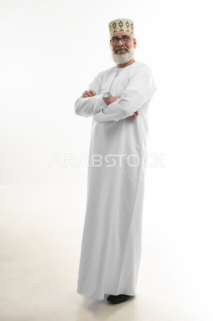 الوقوف بيدين متقاطعتين بإيماءات الثقة بالنفس، الاهتمام بالمظهر الخارجي، مفهوم الرجولة والوقار، بورتريه لرجل كبير بالسن عربي خليجي عماني يرتدي الدشداشة التقليدية والكمه العمانية والنظارة الطبية ينظر إلى الأعلى بتركيز، الجسم بالطول الكامل، خلفية بيضاء