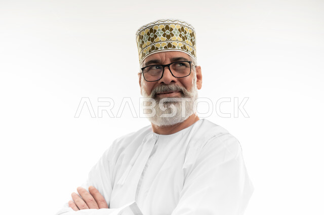 Looking up with gestures of self-confidence, concern for appearance, concept of masculinity and dignity, close-up portrait of an elderly Arab Gulf Omani man wearing the traditional dishdasha, Omani kummah and eyeglasses standing with his hands folded, white background
