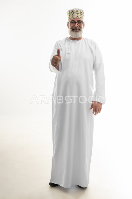 تلقي الاستحسان بشكل كبير، الوقوف والنظر إلى الكاميرا بإيماءات السعادة والسرور، بورتريه لرجل شايب عربي خليجي عماني يرتدي الدشداشة والكمه العمانية يرفع اصبع الإبهام نحو الأعلى، علامة الجودة والقبول، الجسم بالطول الكامل، خلفية بيضاء