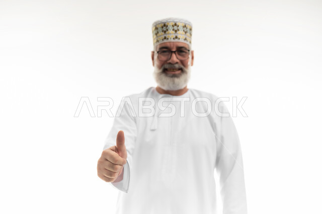 النظر إلى الكاميرا بإيماءات السعادة والسرور، تلقي الاستحسان بشكل كبير، بورتريه مقرب لرجل شايب عربي خليجي عماني يرتدي الدشداشة والكمه العمانية يرفع اصبع الإبهام نحو الأعلى، علامة الجودة والقبول، خلفية بيضاء