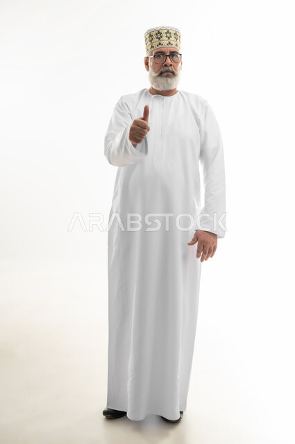 النظر إلى الكاميرا بإيماءات الغضب والانزعاج، بورتريه لرجل شايب عربي خليجي عماني يرتدي الدشداشة والكمه العمانية يرفع اصبع الإبهام نحو الأعلى، علامة الجودة والقبول، تلقي الاستحسان مع عدم الرضا بشكل كامل، الجسم بالطول الكامل، خلفية بيضاء