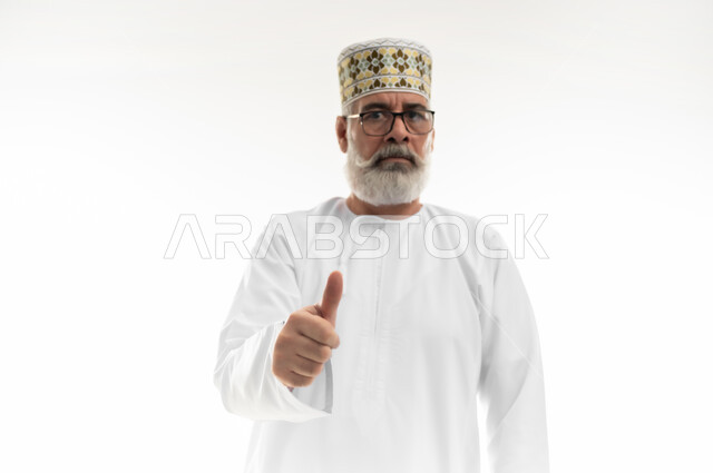 تلقي الاستحسان مع عدم الرضا بشكل كامل، النظر إلى الكاميرا بإيماءات الغضب والانزعاج، بورتريه مقرب لرجل شايب عربي خليجي عماني يرتدي الدشداشة والكمه العمانية يرفع اصبع الإبهام نحو الأعلى، علامة الجودة والقبول، خلفية بيضاء