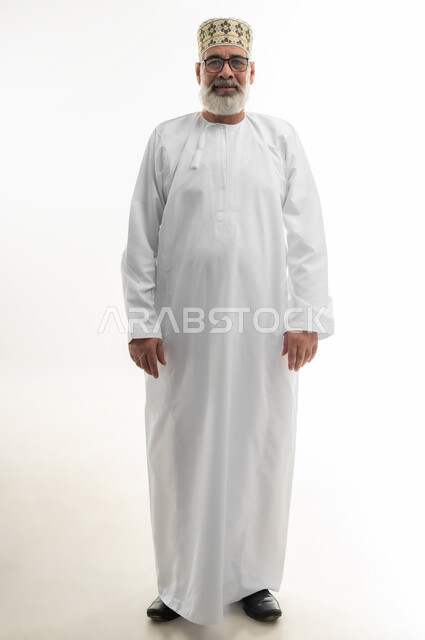 مفهوم الرجولة والوقار، الاهتمام بالمظهر الخارجي، بورتريه لرجل كبير بالسن عربي خليجي عماني يرتدي الدشداشة التقليدية والكمه العمانية والنظارة الطبية يقف بشكل مستقيم وينظر إلى الكاميرا بإيماءات البشاشة والسرور، الجسم بالطول الكامل، خلفية بيضاء