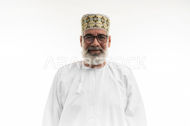 الاهتمام بالمظهر الخارجي، مفهوم الرجولة والوقار، بورتريه مقرب لرجل كبير بالسن عربي خليجي عماني يرتدي الدشداشة التقليدية والكمه العمانية والنظارة الطبية يقف بشكل مستقيم وينظر إلى الكاميرا بإيماءات البشاشة والسرور، خلفية بيضاء