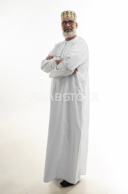 الوقوف بيدين متقاطعتين بإيماءات الثقة بالنفس، الاهتمام بالمظهر الخارجي، مفهوم الرجولة والوقار، بورتريه لرجل كبير بالسن عربي خليجي عماني يرتدي الدشداشة التقليدية والكمه العمانية والنظارة الطبية ينظر إلى مكان ما محدد بتركيز، الجسم بالطول الكامل، خلفية بيضاء