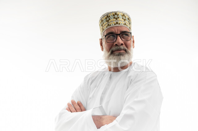 النظر إلى مكان ما محدد بإيماءات الثقة بالنفس، الاهتمام بالمظهر الخارجي، مفهوم الرجولة والوقار، بورتريه مقرب لرجل كبير بالسن عربي خليجي عماني يرتدي الدشداشة التقليدية والكمه العمانية والنظارة الطبية يقف بيدين متكتفتين، خلفية بيضاء