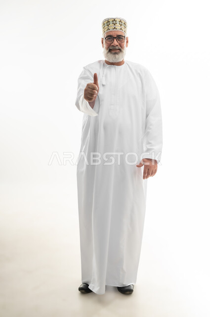 تلقي الاستحسان بشكل كبير، الوقوف والنظر إلى الكاميرا بإيماءات السعادة والسرور، بورتريه لرجل شايب عربي خليجي عماني يرتدي الدشداشة والكمه العمانية يرفع اصبع الإبهام نحو الأعلى، علامة الجودة والقبول، الجسم بالطول الكامل، خلفية بيضاء