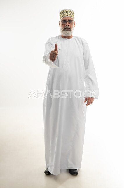 النظر إلى الكاميرا بإيماءات الغضب والانزعاج، بورتريه لرجل شايب عربي خليجي عماني يرتدي الدشداشة والكمه العمانية يرفع اصبع الإبهام نحو الأعلى، علامة الجودة والقبول، تلقي الاستحسان مع عدم الرضا بشكل كامل، الجسم بالطول الكامل، خلفية بيضاء