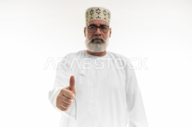 تلقي الاستحسان مع عدم الرضا بشكل كامل، النظر إلى الكاميرا بإيماءات الغضب والانزعاج، بورتريه مقرب لرجل شايب عربي خليجي عماني يرتدي الدشداشة والكمه العمانية يرفع اصبع الإبهام نحو الأعلى، علامة الجودة والقبول، خلفية بيضاء