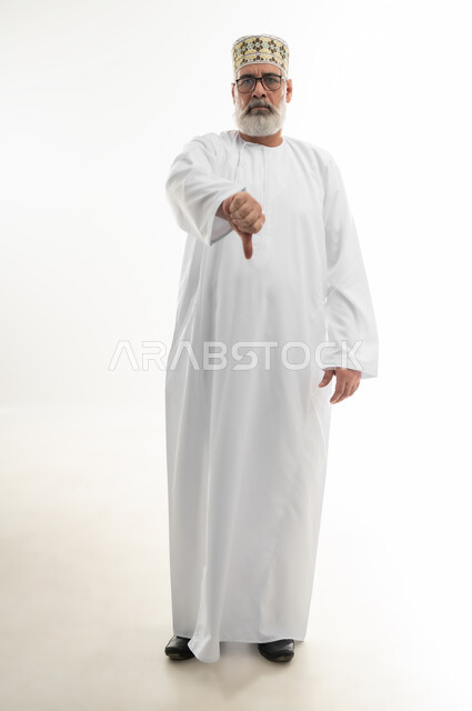 الرفض وعدم القبول، علامات وحركة يد تدل على عدم الإعجاب، النظر إلى الكاميرا بإيماءات الانزعاج والسلبية، بورتريه لرجل شايب عربي خليجي عماني يرتدي الدشداشة والكمه العمانية ويضع النظارة الطبية يشير باصبع الابهام نحو الأسفل، الجسم بالطول الكامل، خلفية بيضاء