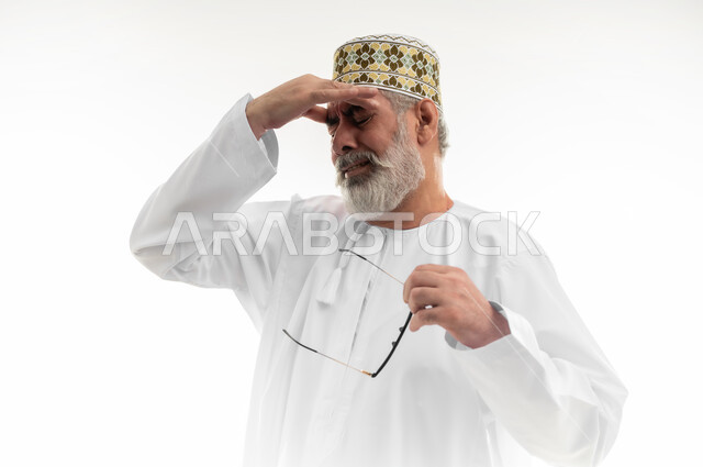 الشعور بالإحباط والخيبة لخسارة شيء ما، إغماض العينين بإيماءات الحزن والرغبة بالبكاء، الأحاسيس والمشاعر المرهفة، بورتريه مقرب لرجل كبير بالسن عربي خليجي عماني يرتدي الدشداشة التقليدية والكمه العمانية ينزع نظارته الطبية ويمسك رأسه، خلفية بيضاء