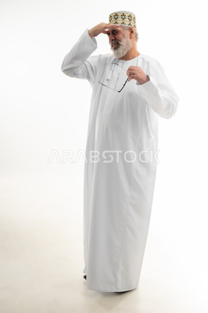 إغماض العينين بإيماءات الحزن والرغبة بالبكاء، الشعور بالإحباط والخيبة لخسارة شيء ما، الأحاسيس والمشاعر المرهفة، بورتريه لرجل كبير بالسن عربي خليجي عماني يرتدي الدشداشة التقليدية والكمه العمانية ينزع نظارته الطبية ويمسك رأسه، الجسم بالطول الكامل، خلفية بيضاء