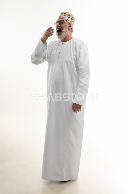 التثاؤب والخمول والحاجة إلى النوم، الإحساس بالتعب والإرهاق وضرورة اخذ قسط من الراحة، بورتريه  لرجل شايب عربي خليجي عماني يرتدي الدشداشة والكمه العمانية والنظارة الطبية يضع يده امام فمه بإيماءات النعاس والكسل، الجسم بالطول الكامل، خلفية بيضاء