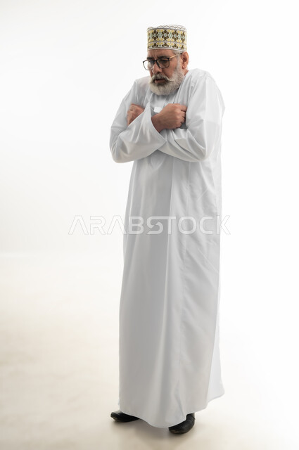 الشعور بالصقيع وتجمد الأطراف، حركات الوجه واليدين تدل على الإحساس بالبرد الشديد، الأجواء الشتوية الباردة، بورتريه لرجل شايب عربي خليجي عماني يرتدي الدشداشة والكمه العمانية التقليدية يحاول تدفئة نفسه، الجسم بالطول الكامل، خلفية بيضاء