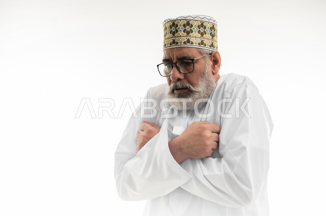 دخول فصل الشتاء وبدء الأجواء الشتوية، الشعور بالصقيع وتجمد الأطراف، حركات الوجه واليدين تدل على الإحساس بالبرد الشديد، بورتريه مقرب لرجل شايب عربي خليجي عماني يرتدي الدشداشة والكمه العمانية التقليدية يحاول تدفئة نفسه، خلفية بيضاء