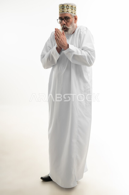 حركات الوجه واليدين تدل على الإحساس بالبرد الشديد، الأجواء الشتوية الباردة، الشعور بالصقيع وتجمد الأطراف، بورتريه لرجل شايب عربي خليجي عماني يرتدي الدشداشة والكمه العمانية التقليدية يحاول تدفئة نفسه، الجسم بالطول الكامل، خلفية بيضاء