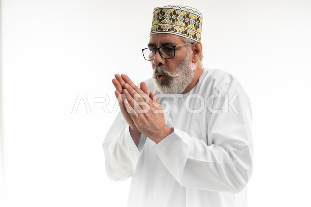 الأجواء الشتوية الباردة، الشعور بالصقيع وتجمد الأطراف، حركات الوجه واليدين تدل على الإحساس بالبرد الشديد، بورتريه مقرب لرجل شايب عربي خليجي عماني يرتدي الدشداشة والكمه العمانية التقليدية يحاول تدفئة نفسه، خلفية بيضاء