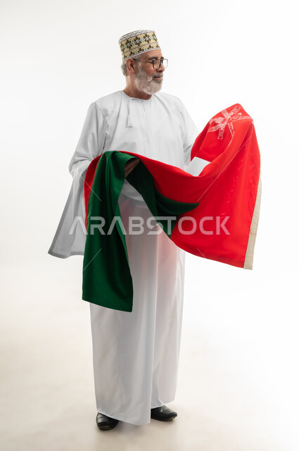 النظر إلى الراية العمانية بمشاعر العز والكرامة، الفخر والاعتزاز بالانتماء لسلطنة عمان، بورتريه لرجل شايب عربي خليجي عماني يرتدي الدشداشة والكمه التقليدية يحمل العلم العماني ويقوم بتقديم الولاء للوطن، الجسم بالطول الكامل، خلفية بيضاء
