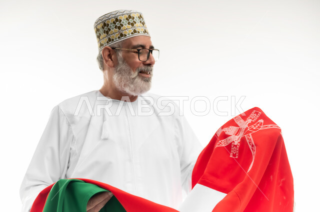 الفخر والاعتزاز بالانتماء لسلطنة عمان، النظر إلى الراية العمانية بمشاعر العز والكرامة، بورتريه مقرب لرجل شايب عربي خليجي عماني يرتدي الدشداشة والكمه التقليدية يحمل العلم العماني ويقوم بتقديم الولاء للوطن، خلفية بيضاء