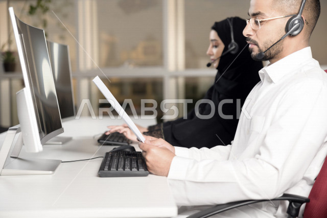موظف عربي خليجي سعودي يعمل في خدمة العملاء ويمسك الاوراق بيده، موظف وموظفة يرتديان سماعة الاتصال، استعلامات واستفسارات، الاجابه على اسئلة العملاء، اجراء اتصال فيديو من خلال الحاسوب، متابعة وتطوير العمل