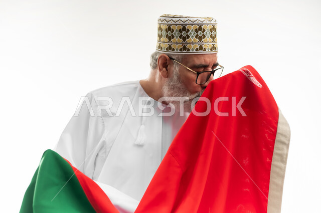 التعبير عن الحب وتقديم الولاء للراية العمانية، الفخر والاعتزاز بالانتماء لسلطنة عمان، بورتريه مقرب لرجل شايب عربي خليجي عماني يرتدي الدشداشة والكمه التقليدية يقبل العلم العماني المغطي لكتفيه بإيماءات العز والكرامة، خلفية بيضاء