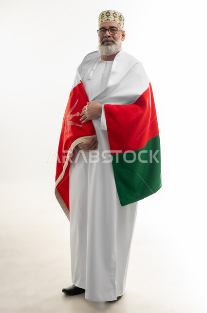 الفخر والاعتزاز بالانتماء لسلطنة عمان، التعبير عن الحب وتقديم الولاء للراية العمانية، بورتريه لرجل شايب عربي خليجي عماني يرتدي الدشداشة والكمه التقليدية يلف العلم العماني على كتفيه وينظر إلى الكاميرا بإيماءات السعادة والسرور، الجسم بالطول الكامل، خلفية بيضاء