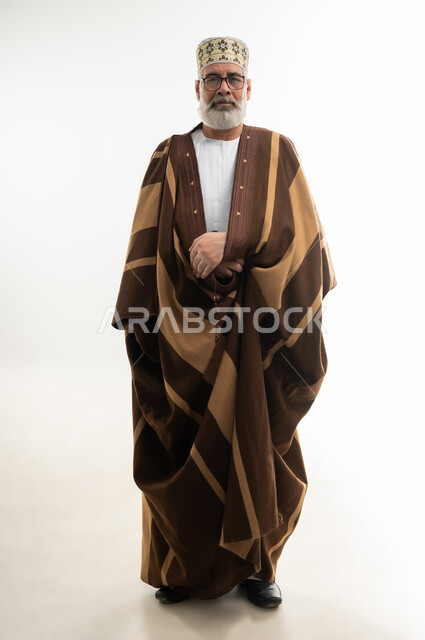 الأجواء الشتوية الباردة، ارتداء الفروة البنية لتدفئة الجسم، مفهوم الهيبة والوقار، بورتريه لرجل شايب عربي خليجي عماني يرتدي الدشداشة والكمه العمانية وينظر إلى الكاميرا بإيماءات البشاشة والسرور، الجسم بالطول الكامل، خلفية بيضاء