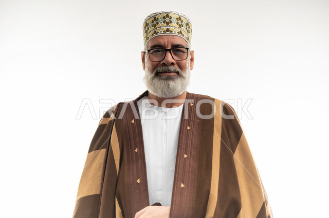 The concept of prestige and dignity, wearing a brown fur coat to warm the body, cold winter atmosphere, close-up portrait of an old Arab Gulf Omani man wearing a dishdasha and an Omani cloak and looking at the camera with gestures of cheerfulness and pleasure, white background