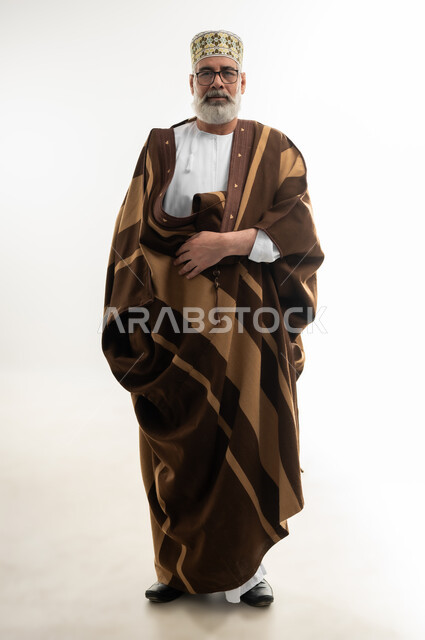 ارتداء الفروة البنية لتدفئة الجسم، الأجواء الشتوية الباردة، مفهوم الهيبة والوقار، بورتريه لرجل شايب عربي خليجي عماني يرتدي الدشداشة والكمه العمانية وينظر إلى الكاميرا بإيماءات البشاشة والسرور، الجسم بالطول الكامل، خلفية بيضاء
