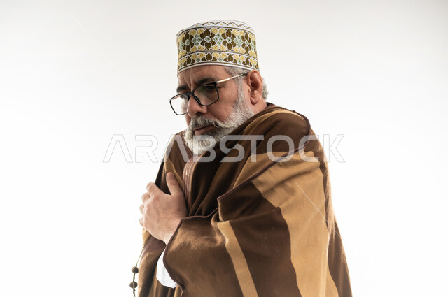 الشعور بالصقيع وتجمد الأطراف، هبوط درجات الحرارة وبدء الأجواء الشتوية، حركات الوجه واليدين تدل على الإحساس بالبرد الشديد، بورتريه مقرب لرجل شايب عربي خليجي عماني يرتدي الدشداشة والكمه والفروة التقليدية في محاولة لتدفئة نفسه، خلفية بيضاء