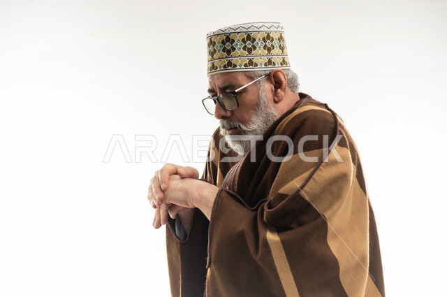 تجمد الأطراف والشعور بالصقيع، هبوط درجات الحرارة وبدء الأجواء الشتوية، حركات الوجه واليدين تدل على الإحساس بالبرد الشديد، بورتريه مقرب لرجل شايب عربي خليجي عماني يرتدي الدشداشة والكمه والفروة التقليدية في محاولة لتدفئة نفسه، خلفية بيضاء