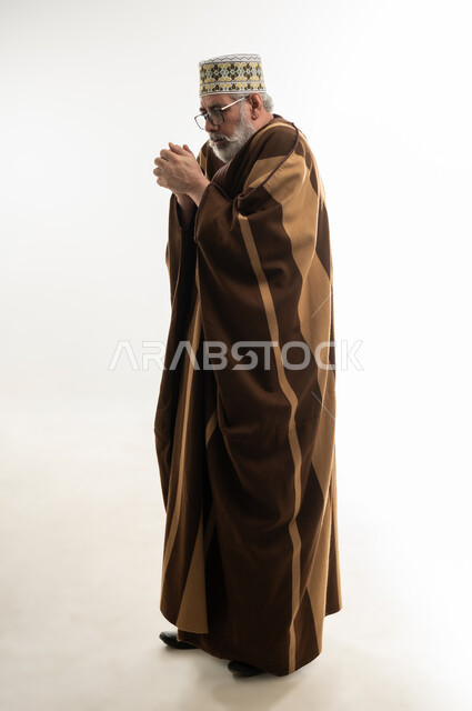 هبوط درجات الحرارة وبدء الأجواء الشتوية، الشعور بالصقيع وتجمد الأطراف، حركات الوجه واليدين تدل على الإحساس بالبرد الشديد، بورتريه لرجل شايب عربي خليجي عماني يرتدي الدشداشة والكمه والفروة التقليدية في محاولة لتدفئة نفسه، الجسم بالطول الكامل، خلفية بيضاء