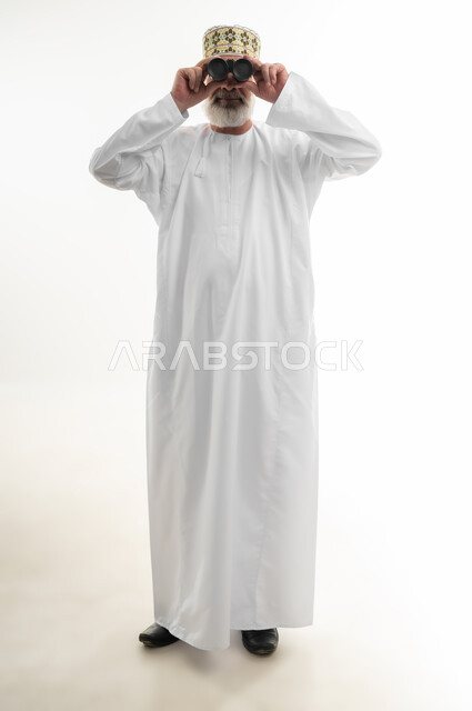 Seeing distant objects using a telescope, love of adventure and exploration, planning for future goals, portrait of an elderly Arab Gulf Omani man wearing the traditional dishdasha and Omani kummah, placing his hand on his forehead and looking through binoculars, full body, white background