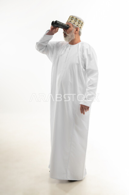 Love of adventure and exploration, seeing distant objects using a telescope, planning for future goals, portrait of an elderly Arab Gulf Omani man wearing the traditional dishdasha and Omani kummah, placing his hand on his forehead and looking through binoculars, full body, white background