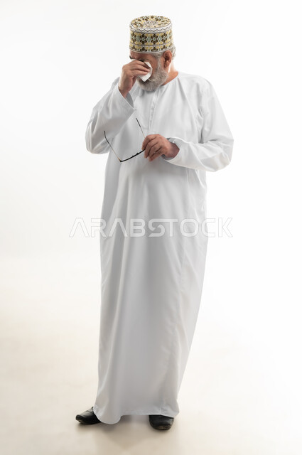 الأحاسيس والمشاعر المرهفة، التأثر بحدث ما والبكاء عليه، الشعور بالحزن والإحباط والخيبة، بورتريه لرجل كبير بالسن عربي خليجي عماني يرتدي الدشداشة التقليدية والكمه العمانية ينزع نظارته ويقوم بمسح دموعه، الجسم بالطول الكامل، خلفية بيضاء