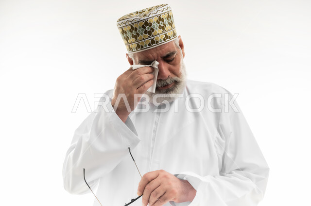 الشعور بالحزن والإحباط والخيبة، الأحاسيس والمشاعر المرهفة، التأثر بحدث ما والبكاء عليه، بورتريه مقرب لرجل كبير بالسن عربي خليجي عماني يرتدي الدشداشة التقليدية والكمه العمانية ينزع نظارته ويقوم بمسح دموعه، خلفية بيضاء