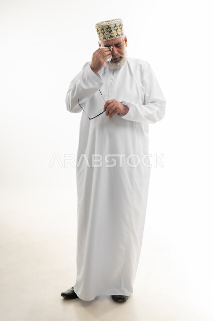 التأثر بحدث ما والبكاء عليه، الأحاسيس والمشاعر المرهفة، الشعور بالحزن والإحباط والخيبة، بورتريه لرجل كبير بالسن عربي خليجي عماني يرتدي الدشداشة التقليدية والكمه العمانية ينزع نظارته ويقوم بمسح دموعه، الجسم بالطول الكامل، خلفية بيضاء