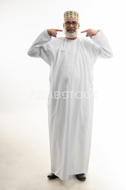 الاهتمام بالمظهر الخارجي، الإشارة إلى النفس بايماءات الثقة، بورتريه لرجل كبير بالسن عربي خليجي عماني يرتدي الدشداشة التقليدية والكمه العمانية يضع يده فوق جبينه وينظر إلى الكاميرا بتعابير البشاشة والسرور، الجسم بالطول الكامل، خلفية بيضاء